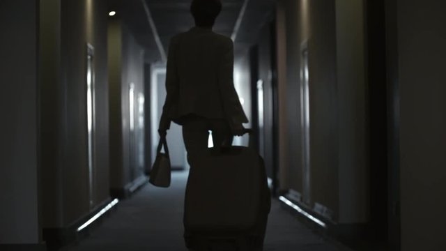 Rear view of silhouette of businesswoman walking with luggage through dark corridor in hotel