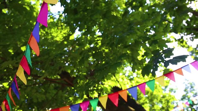 Colorful cute triangle flags hanging on branches of trees as festive decoration for outdoor party. Bright summer sunshine among green leaves in background. Real time full hd video footage.