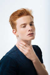 Humourless. Attractive unsmiling red-haired young man touching his neck and wearing a black T-shirt and thinking