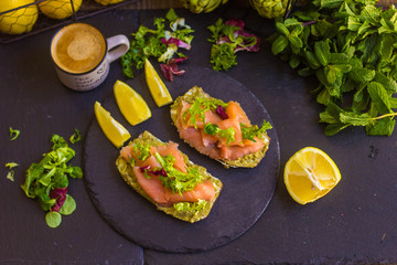 morning breakfast with hot coffee and a sandwich with avocado, soft cheese, lemon and red fish trout on a dark background