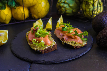 morning breakfast with hot coffee and a sandwich with avocado, soft cheese, lemon and red fish trout on a dark background