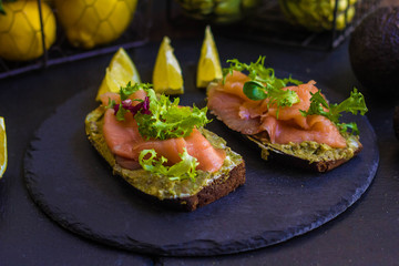 morning breakfast with hot coffee and a sandwich with avocado, soft cheese, lemon and red fish trout on a dark background