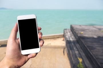 Mockup image of a hand holding and showing white mobile phone with blank black desktop screen nearby the sea and old wooden balcony and blue sky background