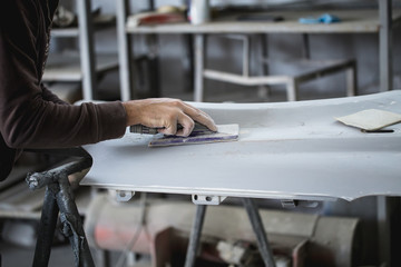 Car detailing - Man with sandpaper in auto repair shop sanding polishing and preparing car parts for painting. Selective focus on man's hand.