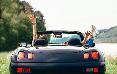 Two girls travelers sit in cabriolet car and enjoy with beautiful view - obrazy, fototapety, plakaty