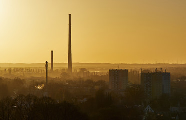 Skyscrapers and chimneys of the factory among trees, buildings and fog.