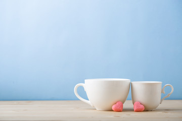 Valentines day concept, mugs cups of coffee or tea for two lovers honeymoon wedding morning surprise breakfast, pink heart sugar candies, blue background pastel colors copy space, light wooden table 