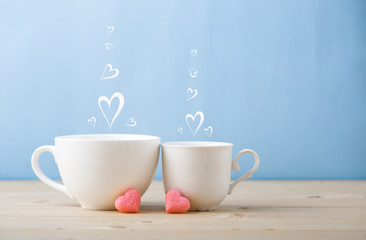 Valentines day concept, mugs cups of coffee or tea for two lovers honeymoon wedding morning surprise breakfast, pink heart sugar candies, blue background pastel colors copy space, light wooden table 