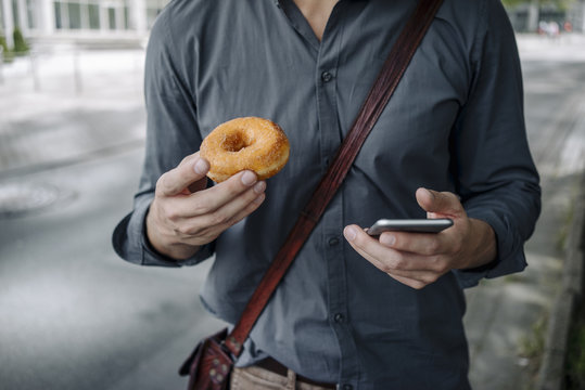 Hands of businessman holding doughnut and smartphone, Partial view - Powered by Adobe