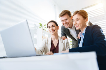 Fototapeta premium Business success. Gay vigorous three colleagues smiling while staring at the screen and man putting hands on women shoulders