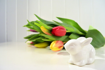 Spring easter composition with fresh tulips and porcelain bunny on white table. Wooden planks background