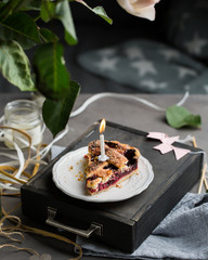 A piece of a birthday cherry cake with a burning one white candle. Atmospheric beautiful food birthday photo with home cherry pie, flowers, ribbons and decoration.