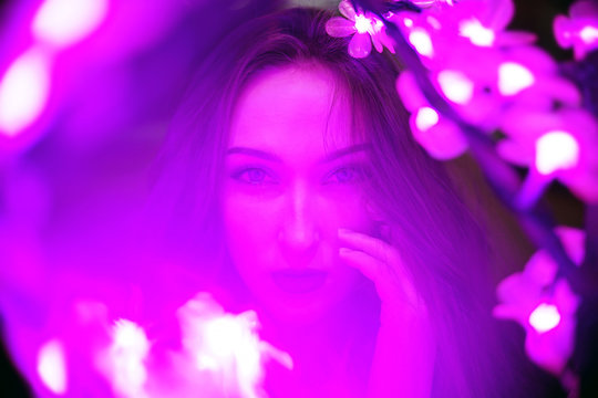 Sexy young beautiful woman posing near neon sakura tree lights dramatic ultraviolet background