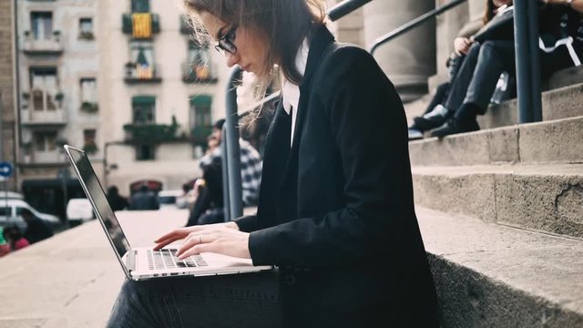 Close-up of young female student sitting on the stairs and typing on computer keyboard, looking serious and concentrated, outdoors, slow motion