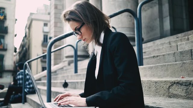 Side view of young attractive businesswoman using modern laptop outdoors, female student typing on computer keyboard while sitting on the stairs of old historical building, slow motion