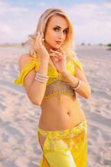 A beautiful blond girl in a yellow dress, suit is dancing an oriental, East dance in the desert on the sand. Beautiful exotic woman dancer in a costume for belly dancing.