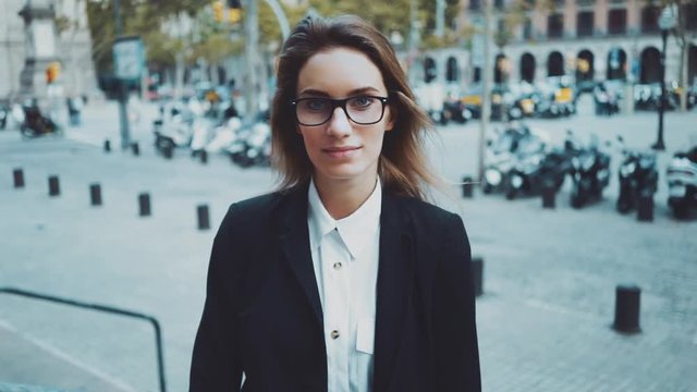 Portrait of young smiling businesswoman looking at camera outdoors, female student wearing glasses and looking at camera with smile, slow motion