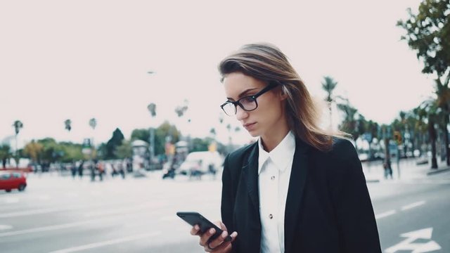 Professional young businesswoman using modern smartphone while going to her office on urban street, slow motion