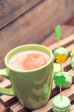 Hot cocoa in green cup and green macaroon cookies scattered on the wooden surface with St. Patrick’s Day attributes. Tinted photo. Shallow depth of field. Copy space