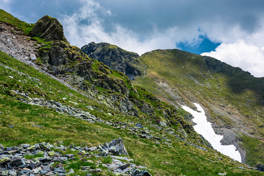 rocky cliffs on grassy slopes with snow in summer. lovely nature scenery under the cloudy sky in Fagaras mountains, Romania