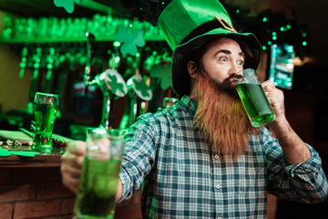 A man in a leprechaun hat and with a beard drinks beer in a bar.