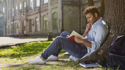 University student sitting under tree reading book with gripping plot, engrossed