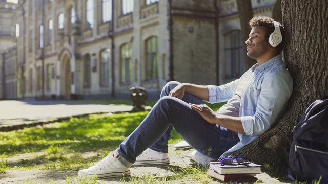 Mixed Young Male Sitting Under Tree In Headphones, Eyes Closed, Enjoying Music