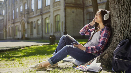 Female student sitting on grass, leaning against tree, listening to music, relax