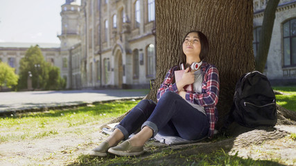 Mixed female reading book under tree, pressing it against chest, favorite book