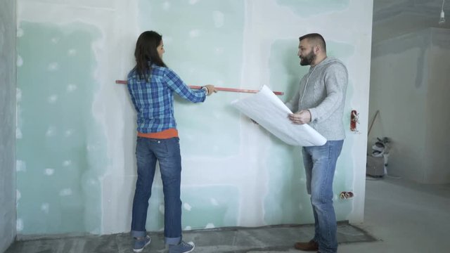 Couple with blueprints checking wall with level at their new home 
