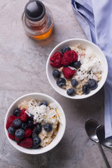 Overhead shot of oatmeal served with berries and coconut . Spoons, maple syrup and a blue napkin next to the bowls. 