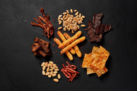 Top View Of Various Beer Snacks Like Peanut, Crackers, Sausages, Chips, Jerky, Cheese Sticks On Dark Desk.