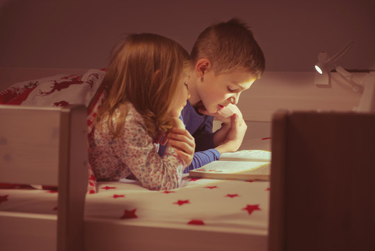 Two happy sibling children reading book in bunk bed under blanket