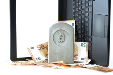 stone monument/tombstone with bitcoin symbol standing on a pile of banknotes in front of a flipped black notebook on white background - copy space for text on display