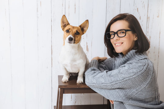 Photo of adorable glad satisfied female wears spectacles and loose warm sweater, stands near her favourite dog who sits on chair, look at camera, going to have walk outdoor. People and pets.