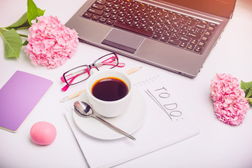 Day Planning concept. Cup of coffee on notebook with To do list, laptop, sketchbook, glasses, and wisteria flowers on the white working office desk. Selective focus.