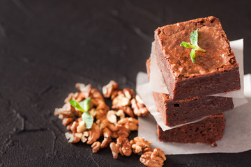 Chocolate brownie square pieces in stack on white plate with walnuts, decorated with mint leaves and cocoa on black background. Delicious dessert. Dark mood. Close up photography. Selective focus