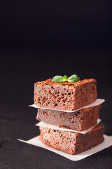 Chocolate brownie square pieces in stack on white plate with walnuts, decorated with mint leaves and cocoa on black background. Delicious dessert. Dark mood. Close up photography. Selective focus
