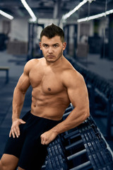Fototapeta premium Muscular bodybuilder in gym near dumbbell rack.Strong athletic man shows body,abdominal muscles,chest muscles,biceps and triceps.Work out,gaining weight. Bodybuilding concept.
