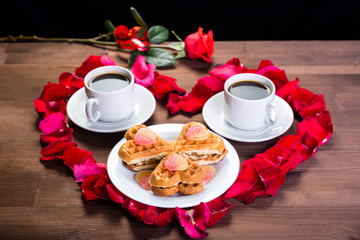 On the wooden table, inside the heart of the rose petals are two cups of coffee and a plate with biscuits. Outside is a rose flower. Front view