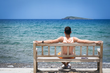 Young man with outstretched arms, sitting on a wooden bench on the shore of the Mediterranean sea on the island of Crete and looking into the horizon