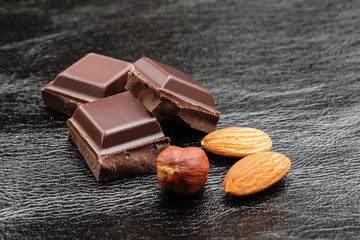 Pieces of bitter dark chocolate cubes with hazelnut and almond nuts on black leather texture background