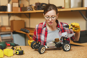 Beautiful caucasian young brown-hair woman in plaid shirt, gray T-shirt, yellow gloves making toy car iron model constructor, working in carpentry workshop at wooden table place with different tools.