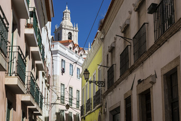 Fototapeta na wymiar View of a narrow street and buildings with a tower of the Sao Vincente de Fora church on the backrgound, in the historic neighborhood of Alfama in Lisbon, Portugal; Concept for visit Lisbon