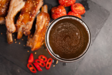 Pork ribs in barbecue sauce and honey baked tomatoes and a glass of beer on a black slate dish. A great snack to beer on a dark stone background. Top view