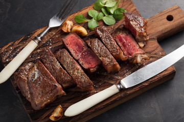 Closeup ready to eat steak new York beef breeds of black Angus with herbs, garlic and butter on a wooden Board. The finished dish for dinner on a dark stone background. Top view
