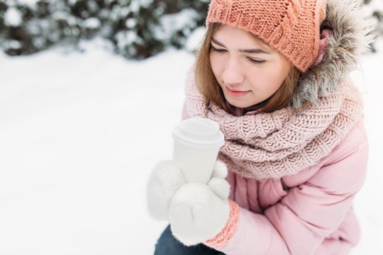 Portrait of a beautiful young girl in white knitted mittens, outdoors, holding a paper Cup with a hot beverage, bright winter day.the woman smiles and is happy, close-up, Trees in first snow in the.