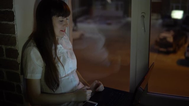 A business woman sits on the windowsill and waves her hand across the laptop during a video conference evening, outside the window in the street a road with a busy traffic.