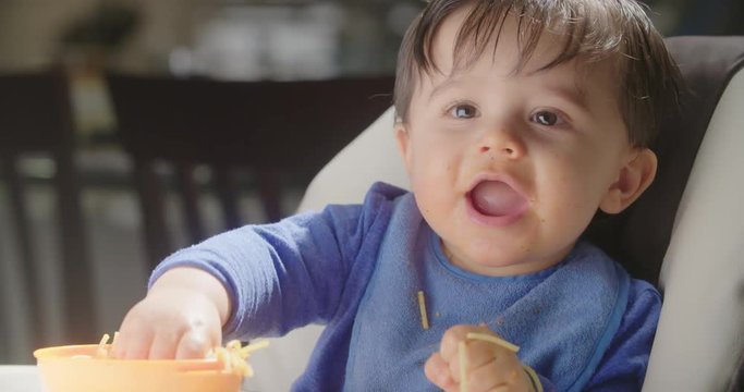 Baby Plays with Food in Highchair and Laughs. a medium view of a baby in a highchair playing with his pasta in a bowl. Picking it up and dropping while laughing. Slow Motion