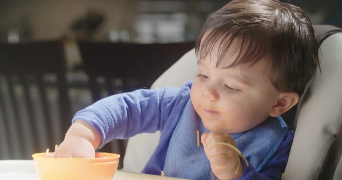 Baby Eats Handful of Pasta Medium. a medium view of a baby in a highchair grabbing a handful of pasta, eating and smiling. Slow motion